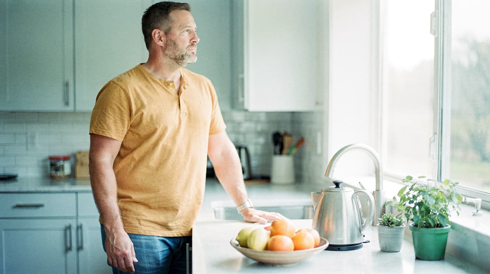 A man in a yellow shirt stands in a kitchen looking out the window, with a bowl of fruit and a kettle on the counter.