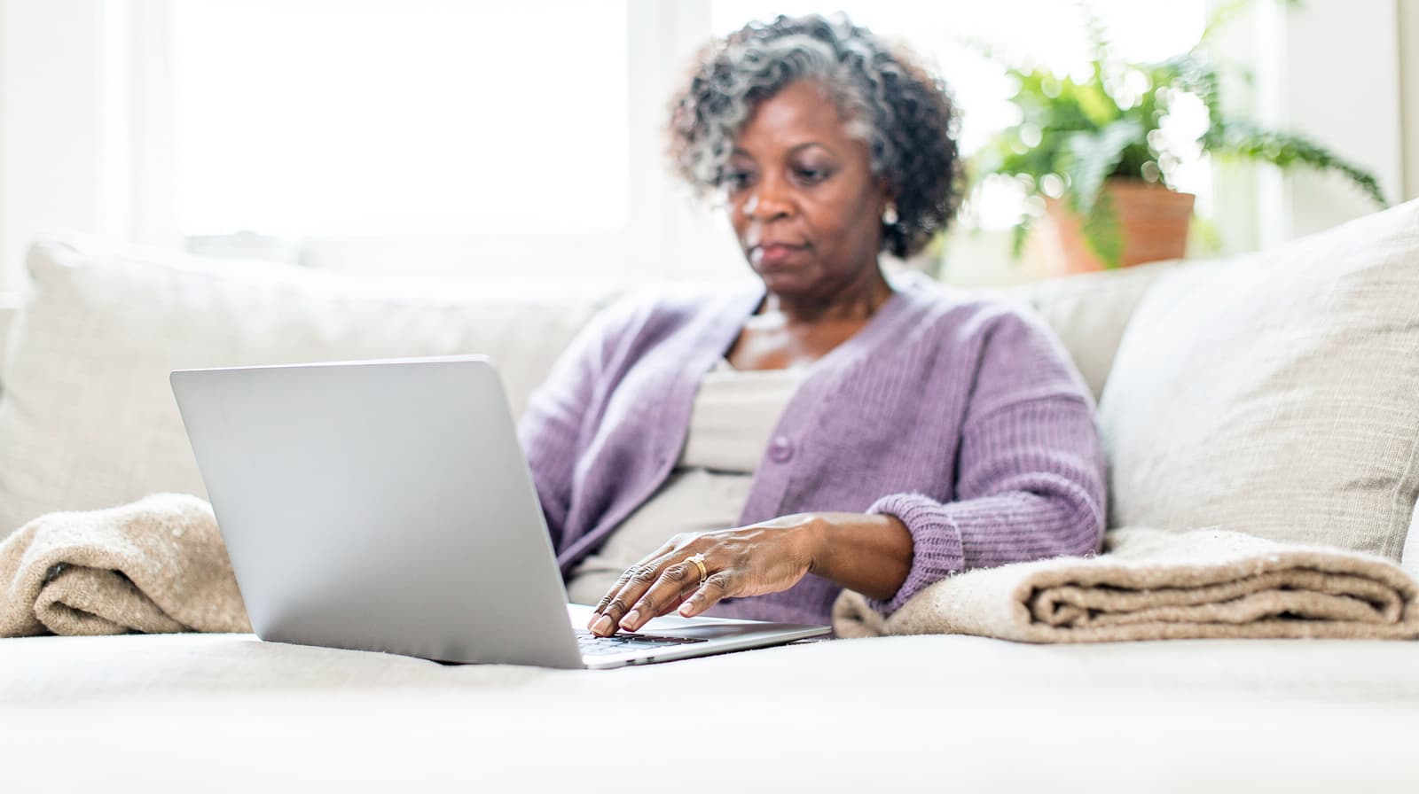 A woman sits on a sofa using a laptop with a plant in the background.