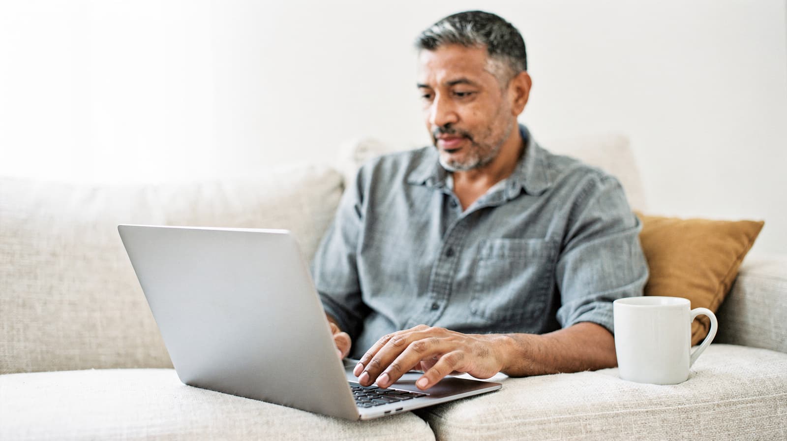 A man sits on a couch using a laptop with a mug beside him.