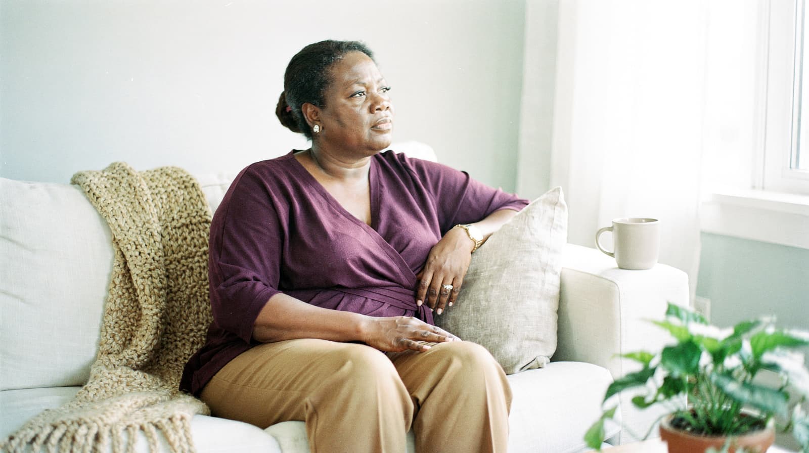 A woman sits on a sofa with a blanket, looking out a window, next to a potted plant and a mug.