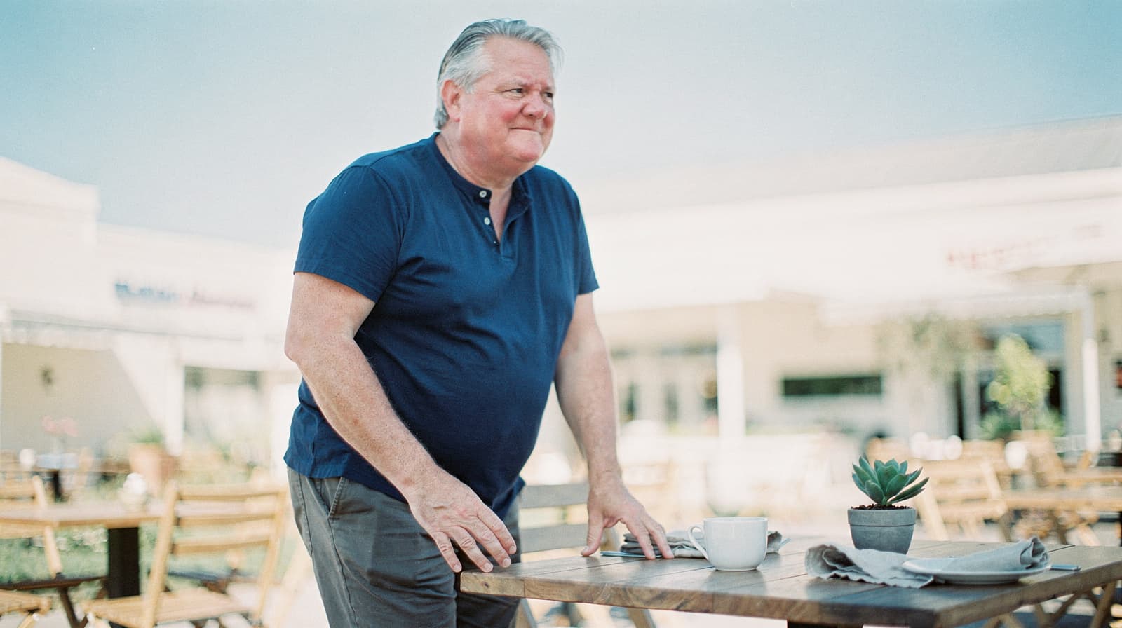 A man in a blue shirt stands by a wooden table with a cup and a small plant in an outdoor seating area.