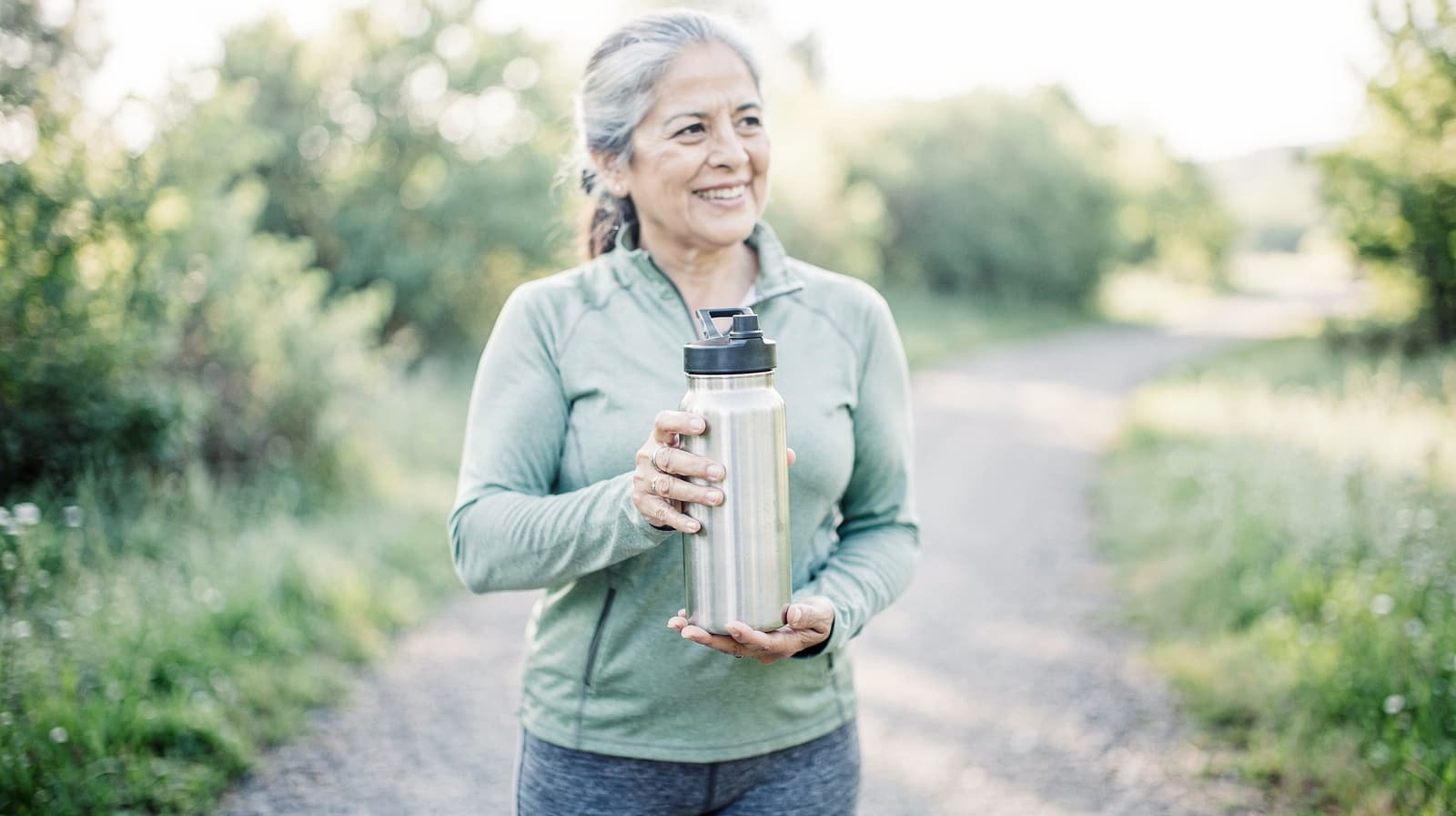 A woman in a green jacket holds a water bottle while standing on a gravel path surrounded by greenery.