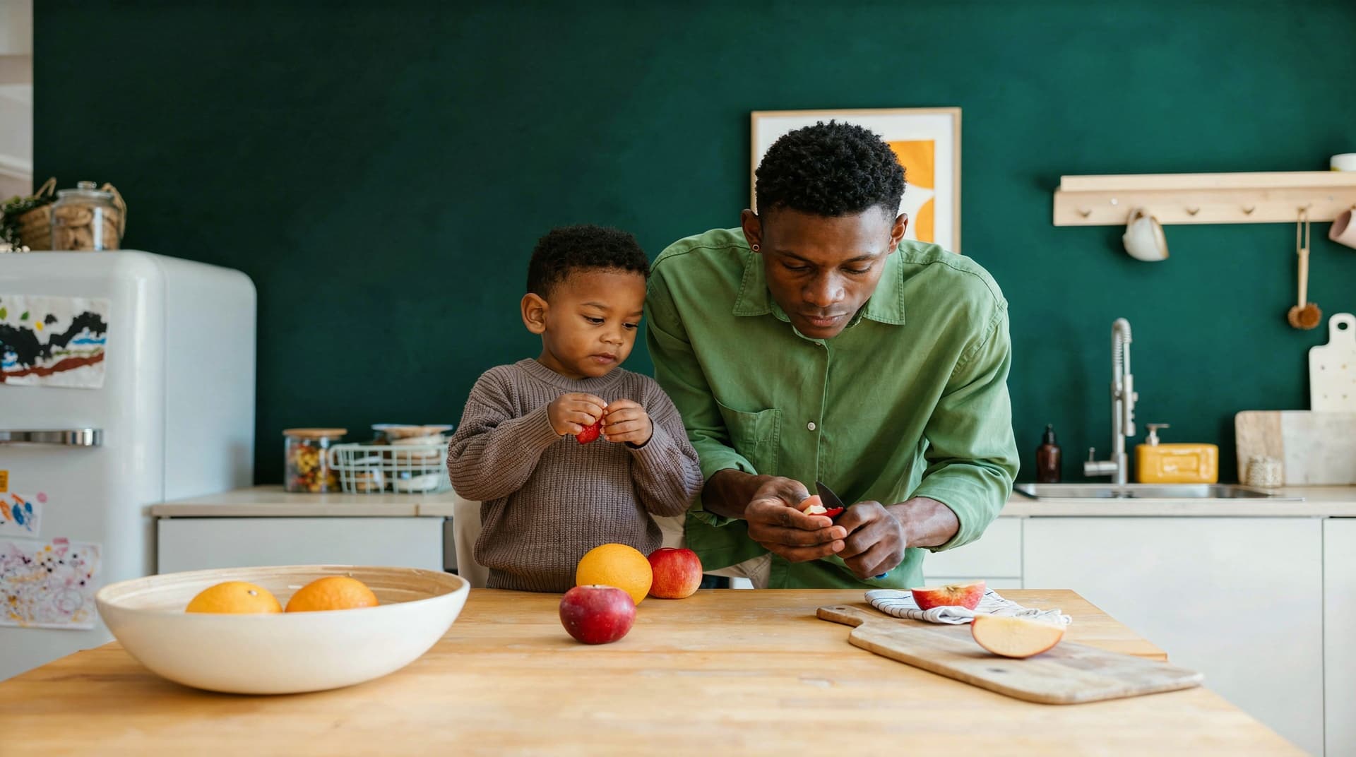 A man and a child peel fruit together at a kitchen counter with a green wall in the background.