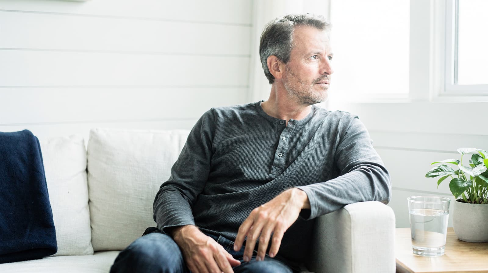 A man sits on a white couch looking out a window with a glass of water and a small plant nearby.