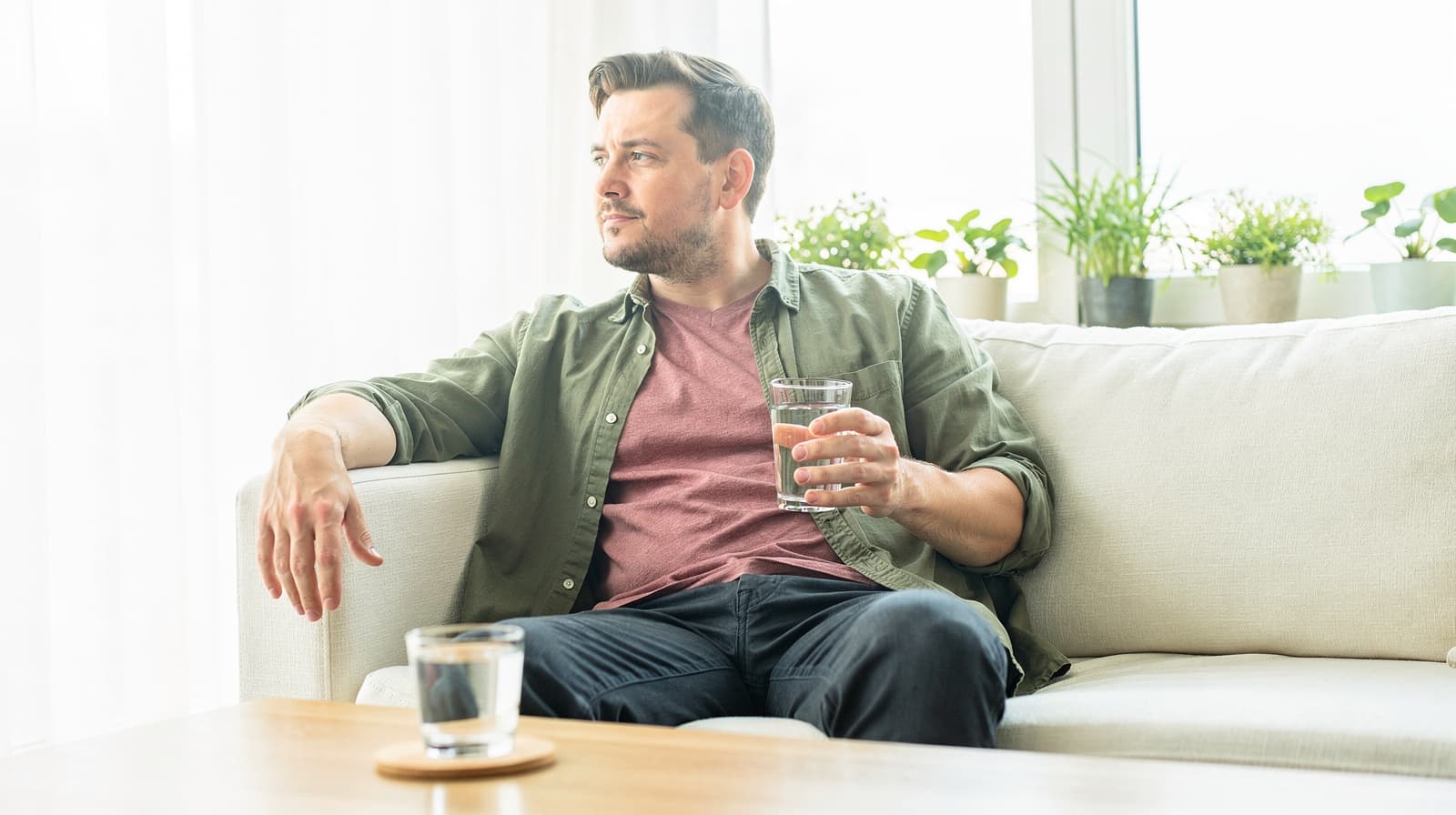 A man sits on a couch holding a glass of water, looking out a window with plants on the windowsill.
