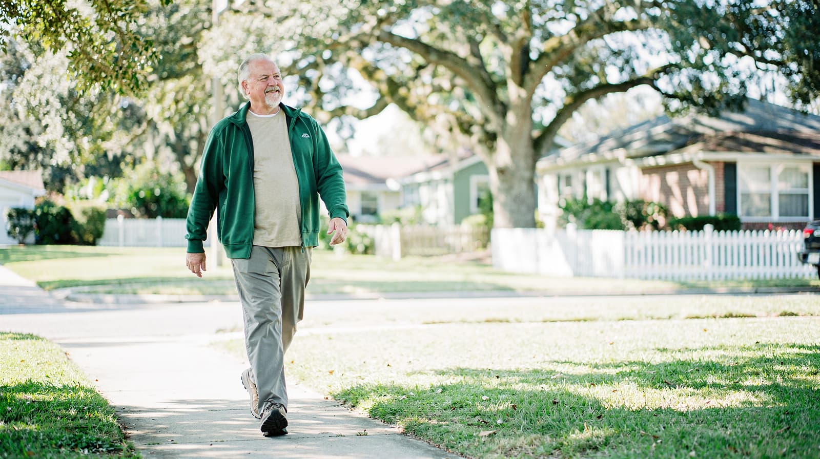 A man in a green jacket walks on a sidewalk in a suburban neighborhood with houses and trees in the background.