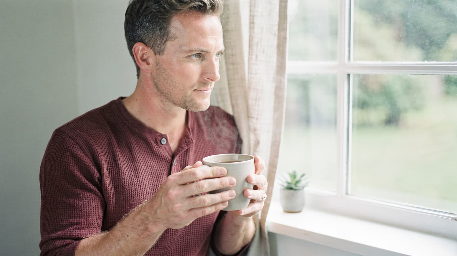 A man in a maroon shirt holds a mug while looking out a window next to a small potted plant.