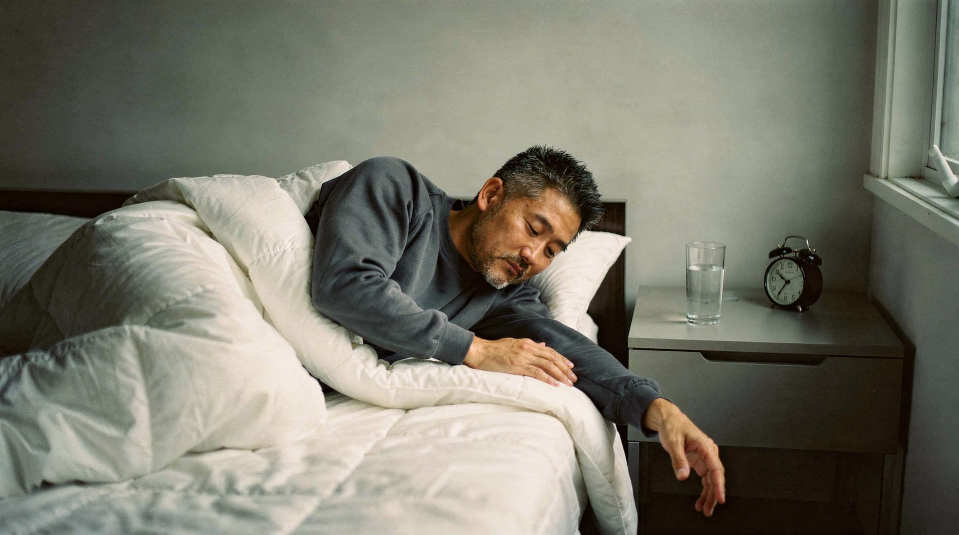 A man lies in bed under a white comforter, reaching toward a nightstand with a glass of water and an alarm clock.