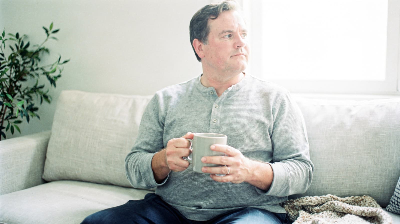 A man sits on a couch holding a mug, looking out a window with a plant nearby.