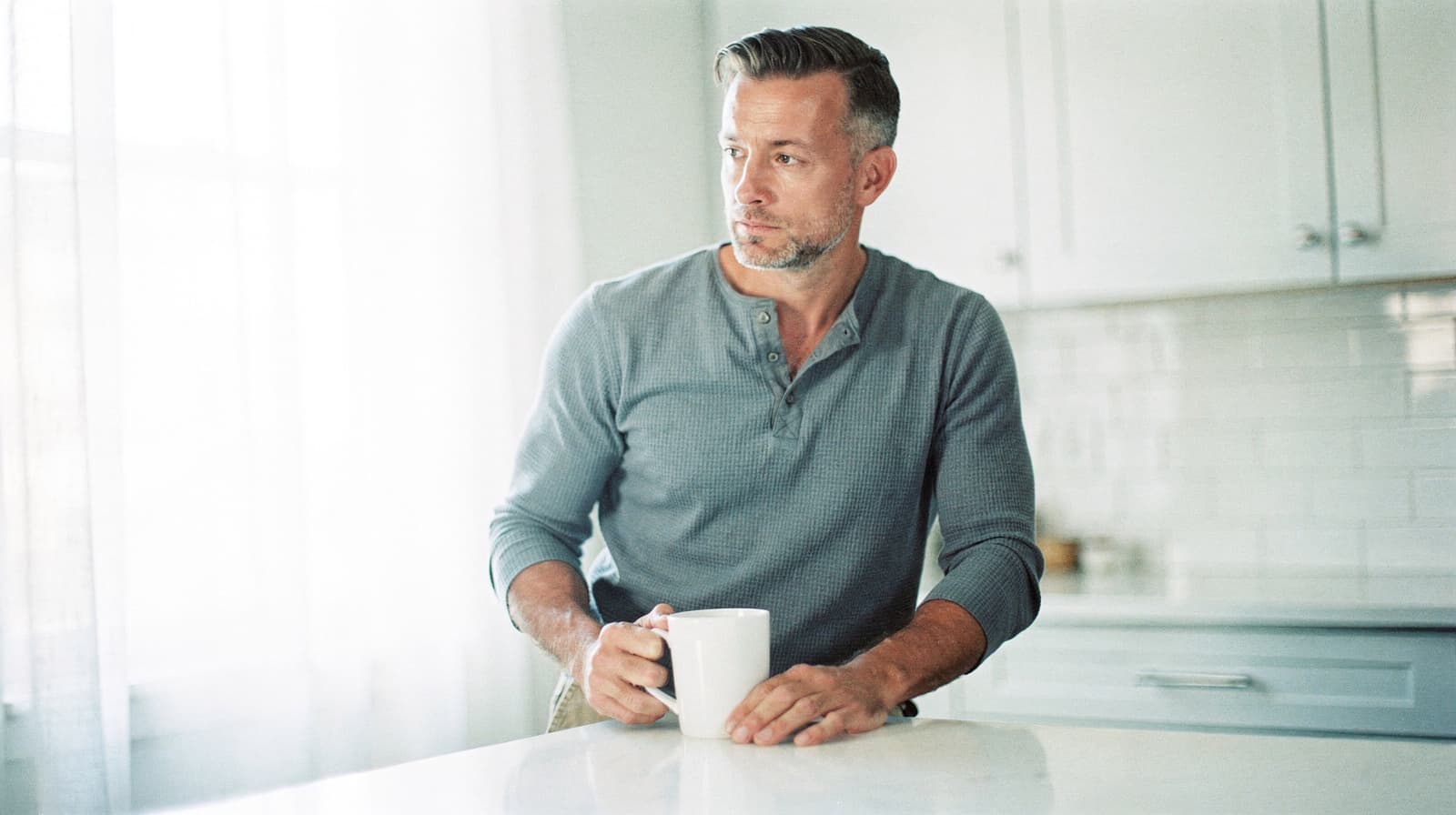 A man in a gray shirt holds a mug while leaning on a kitchen counter with white cabinets in the background.