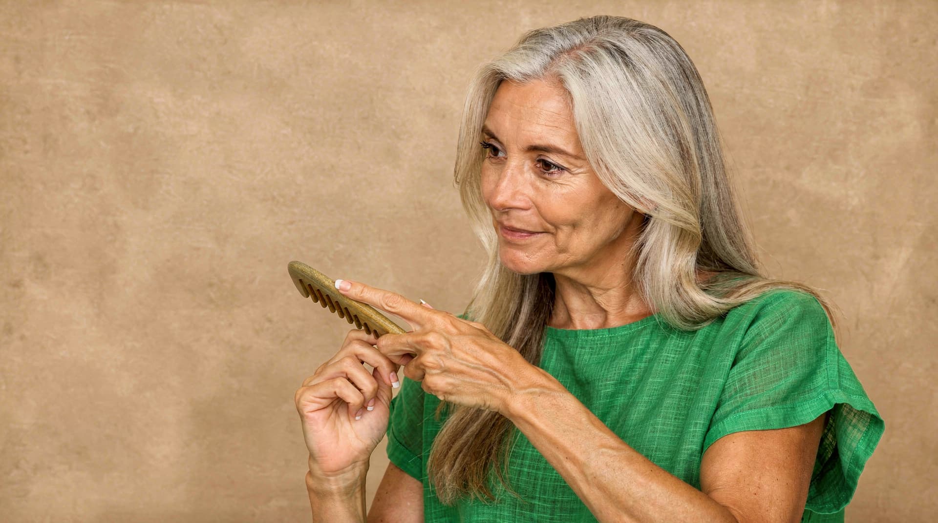 A woman with long gray hair holds a wooden comb while wearing a green shirt against a beige background.