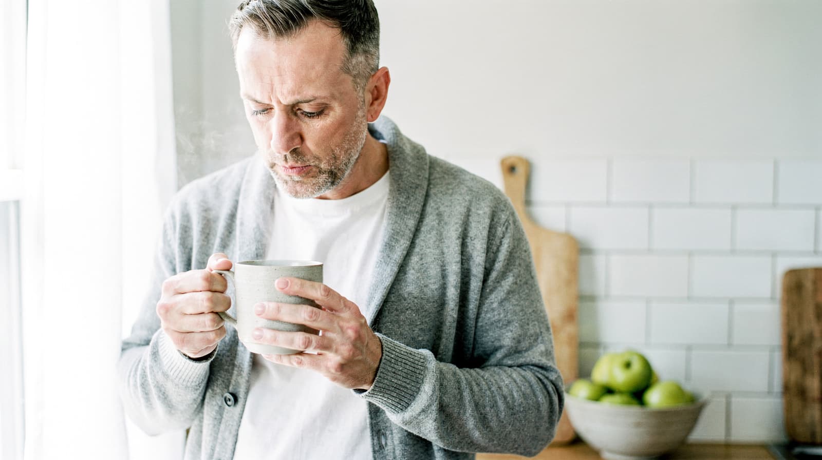 A man in a gray cardigan holds a steaming mug in a kitchen with a bowl of green apples on the counter.