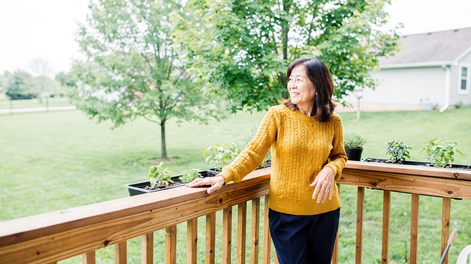 A woman in a yellow sweater stands on a wooden deck, looking out at a grassy yard with trees.