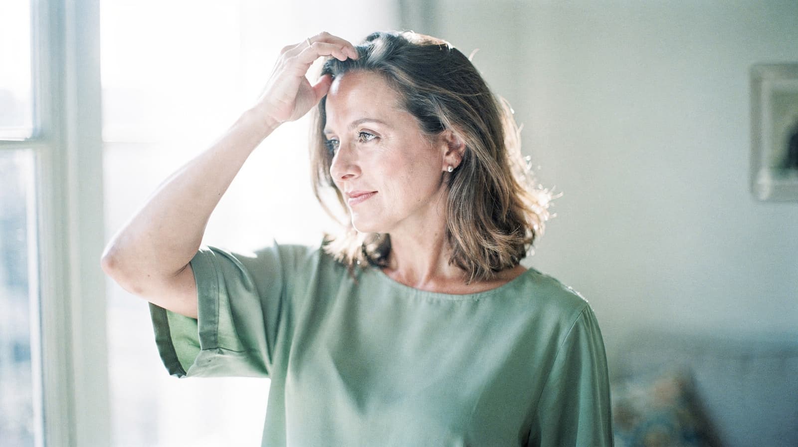 A woman in a green blouse stands indoors, looking out a window with her hand touching her hair.