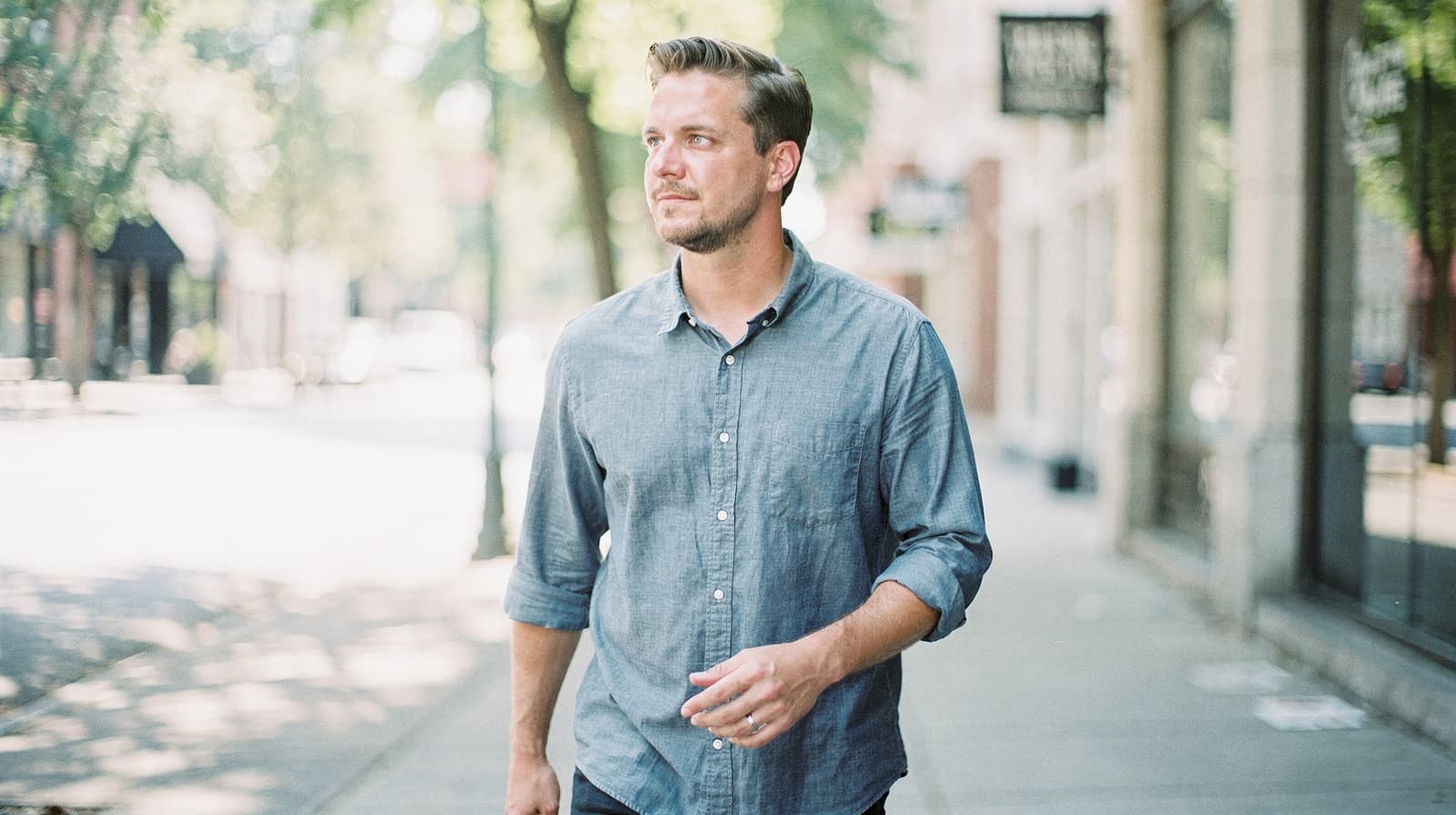 A man in a blue shirt walks along a sunlit city sidewalk lined with trees and shops.