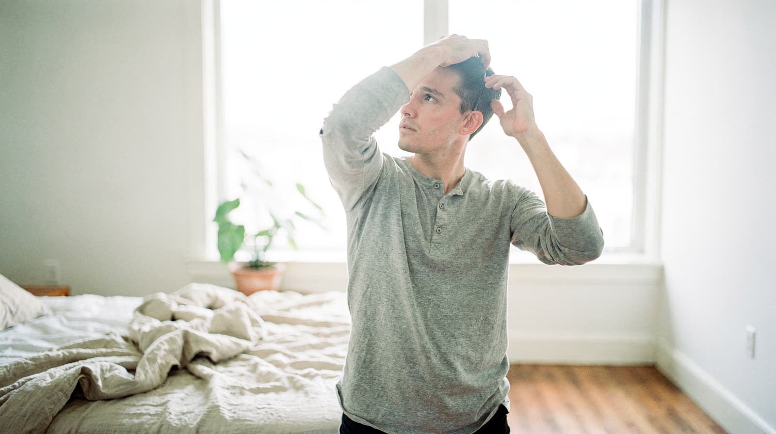 A man in a gray shirt stands in a bedroom adjusting his hair near a bed with rumpled sheets and a potted plant by the window.
