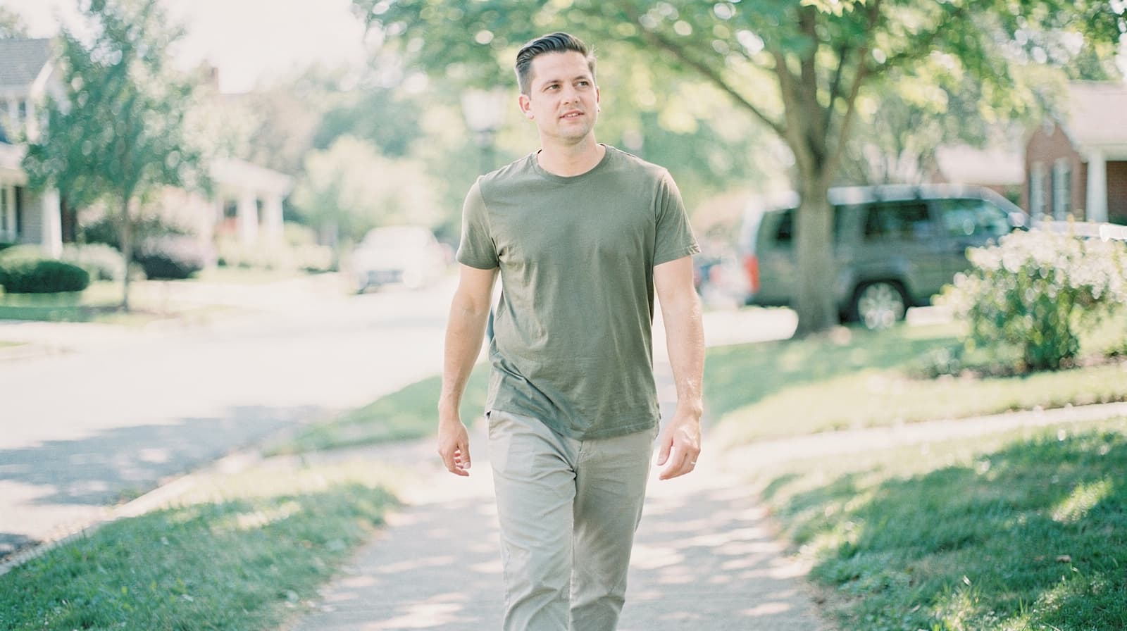 A man in a green shirt walks on a sunny residential sidewalk lined with trees and houses.