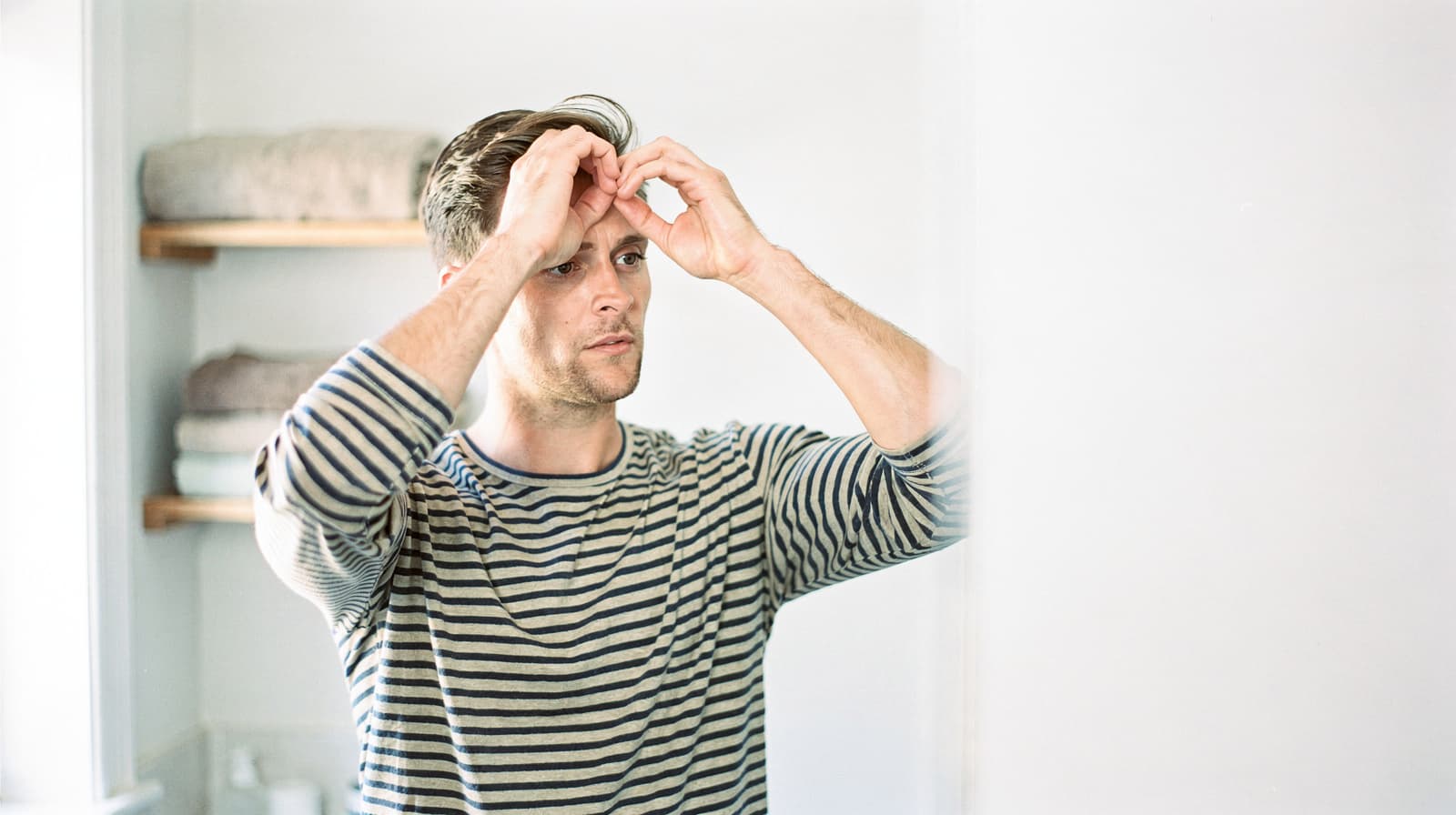 A man in a striped shirt looks at himself in a mirror, adjusting his hair in a bright room with shelves in the background.