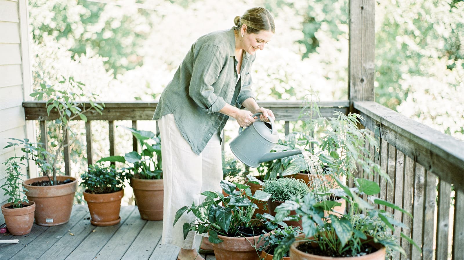 A woman waters potted plants on a wooden porch surrounded by greenery.