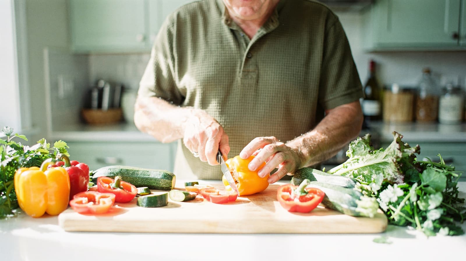 A man in a kitchen slices a yellow bell pepper on a wooden cutting board surrounded by various vegetables.