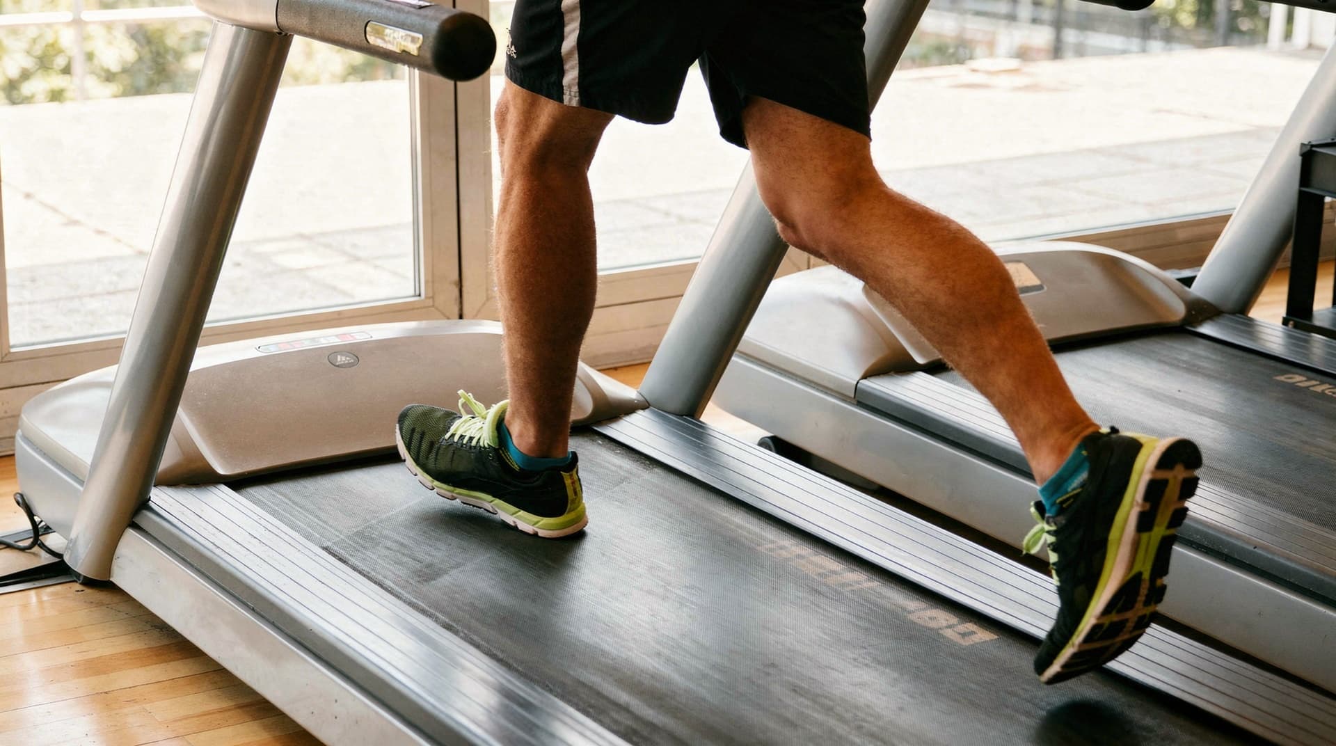 A person in athletic shoes runs on a treadmill in a gym with large windows.
