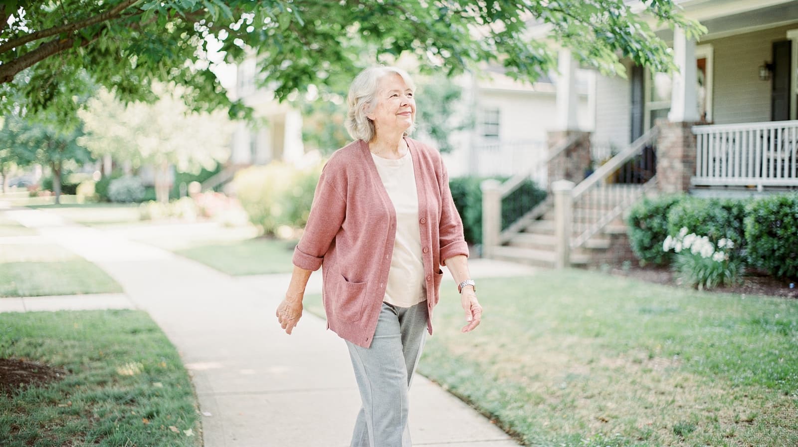 A woman walks along a sidewalk in a residential neighborhood with trees and houses in the background.