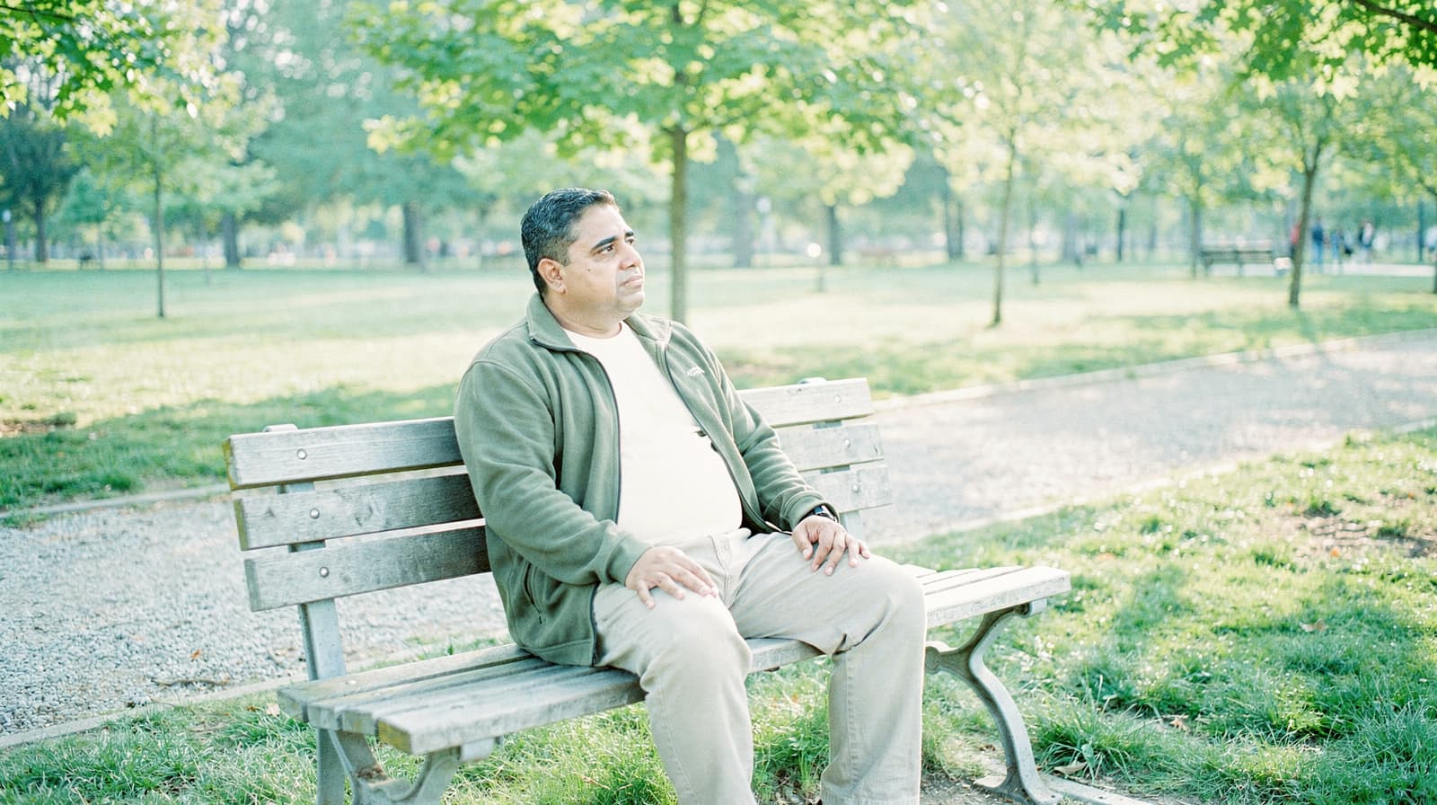 A man sits on a park bench surrounded by trees and grass on a sunny day.