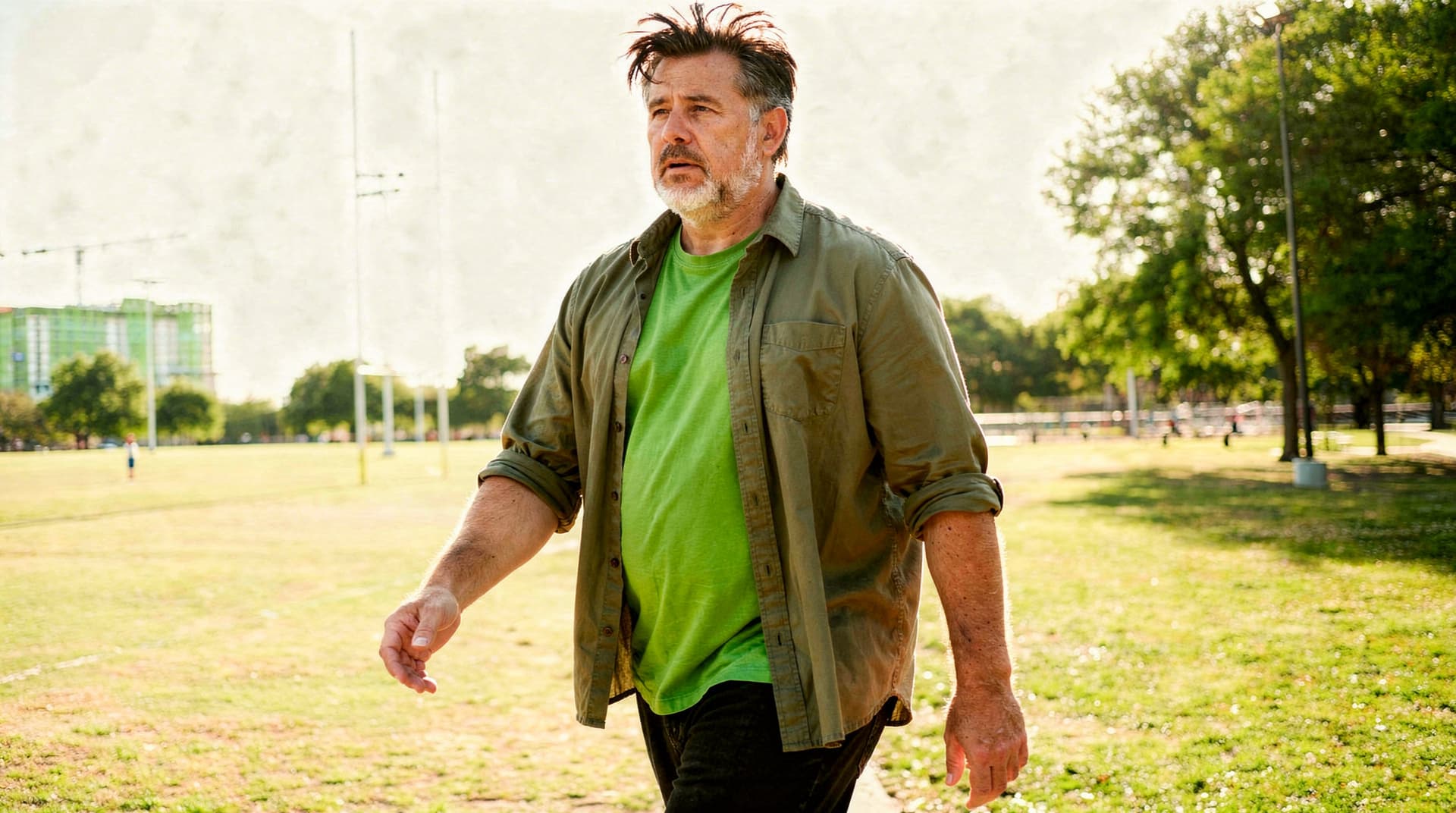 A man in a green shirt walks through a sunny park with trees and a sports field in the background.