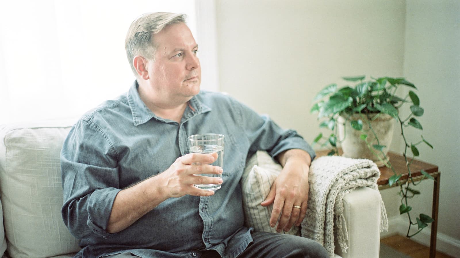 A man sits on a couch holding a glass of water, with a plant on a table nearby.