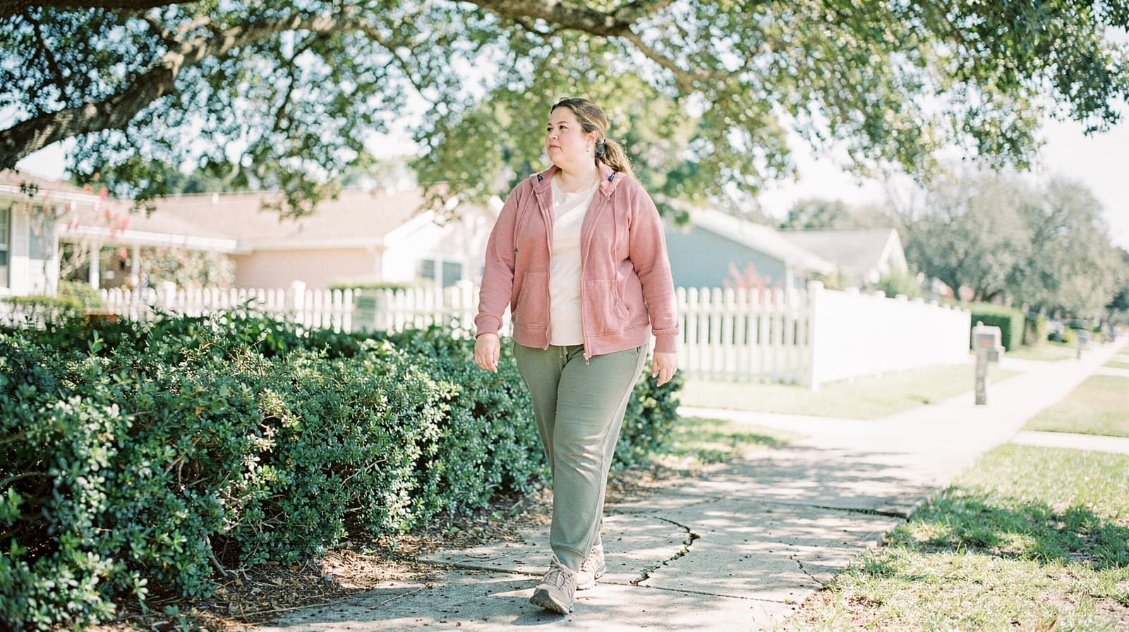 A woman in a pink jacket walks on a sunny sidewalk lined with bushes and trees.