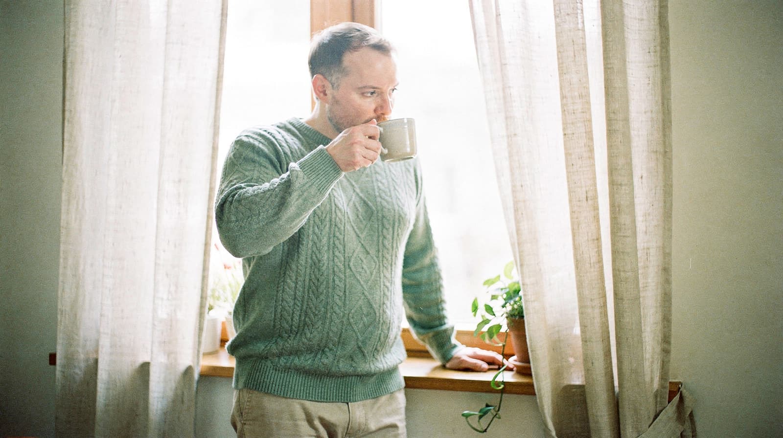 A man in a sweater drinks from a mug while standing by a window with curtains and potted plants.