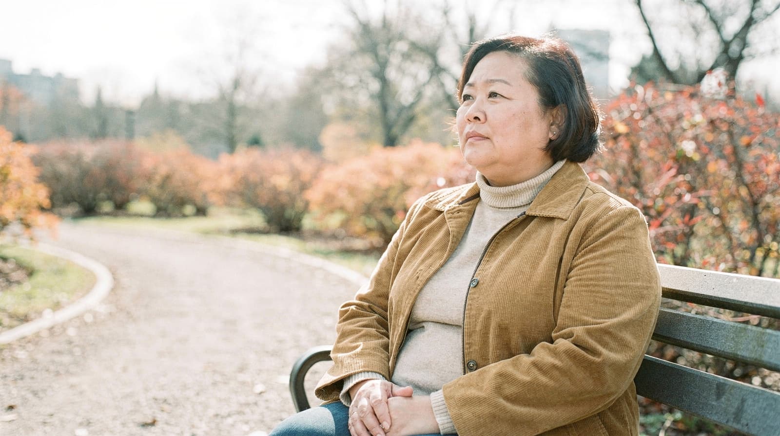 A woman sits on a park bench surrounded by autumn foliage on a sunny day.
