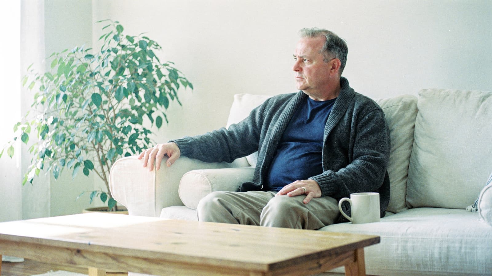 A man sits on a white sofa in a bright room, looking out the window with a mug on the wooden table in front of him.
