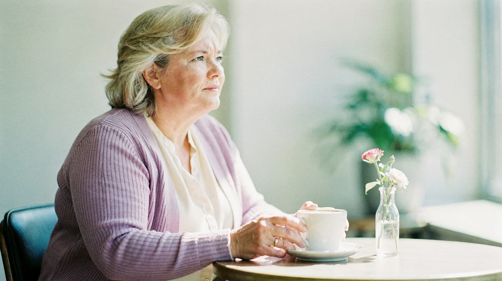 A woman sits at a table holding a cup, with a small vase of flowers nearby.