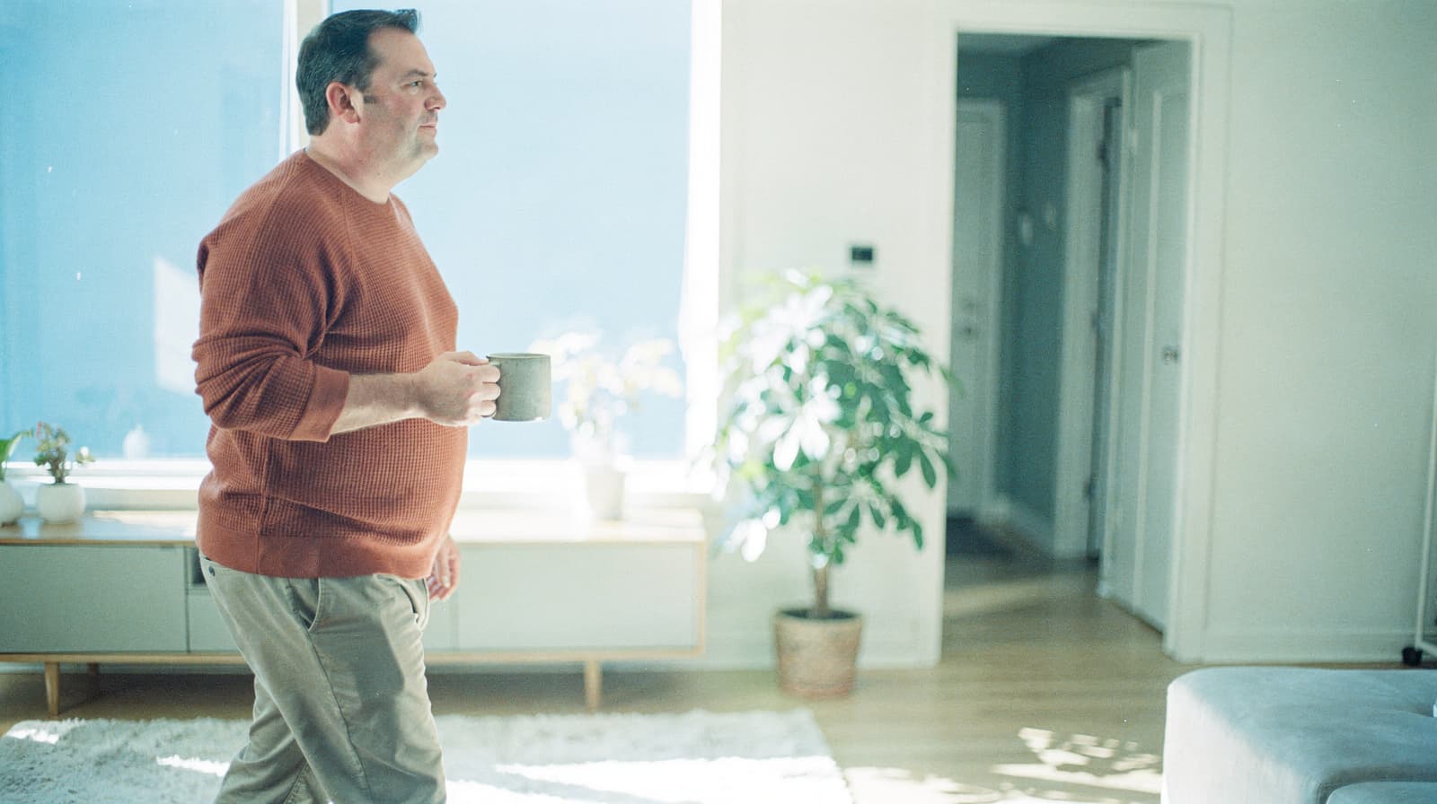 A man holding a mug walks through a sunlit living room with a potted plant and a sideboard.
