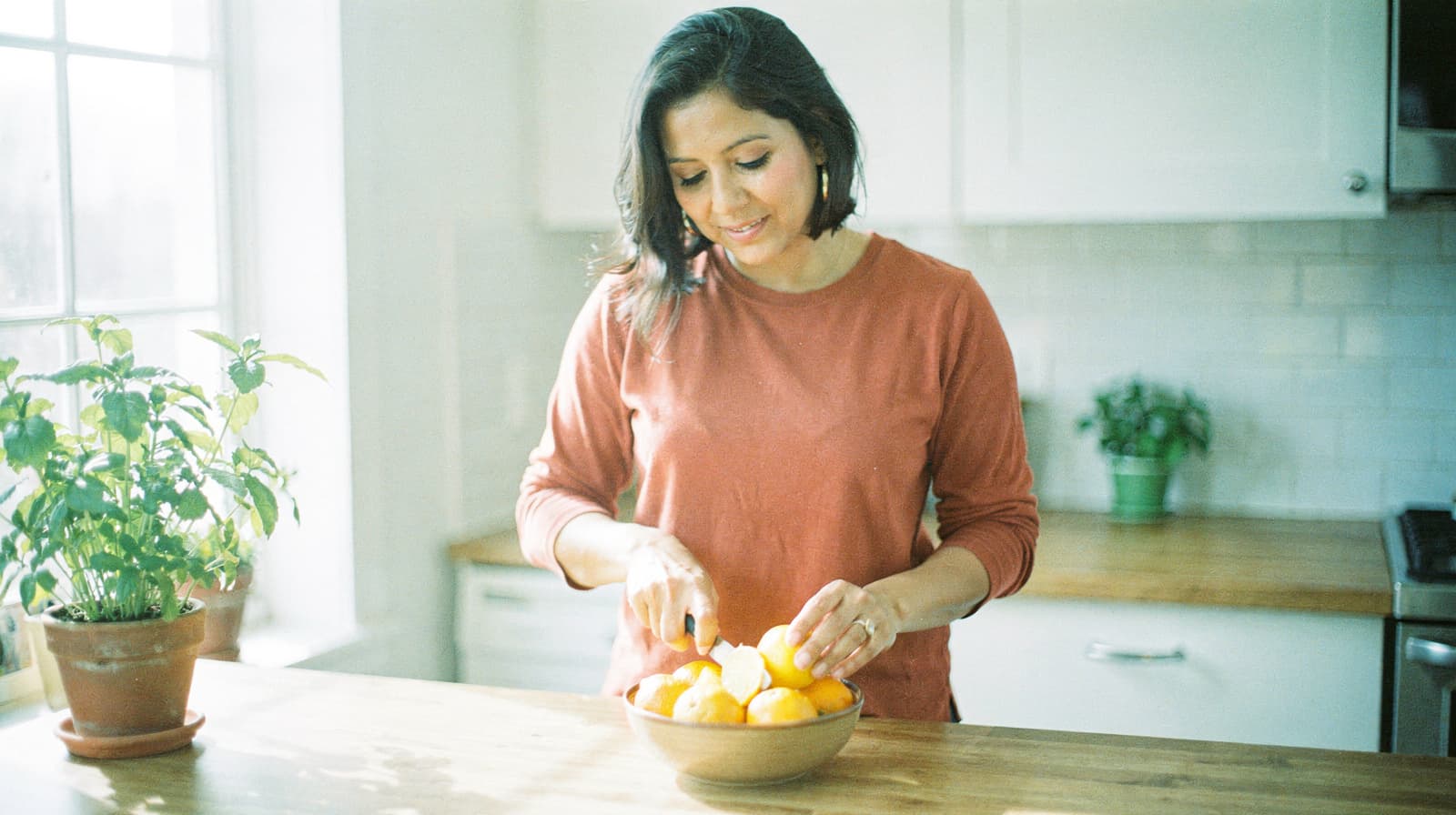 A woman in a kitchen cuts oranges in a bowl on a wooden countertop near a window with potted plants.