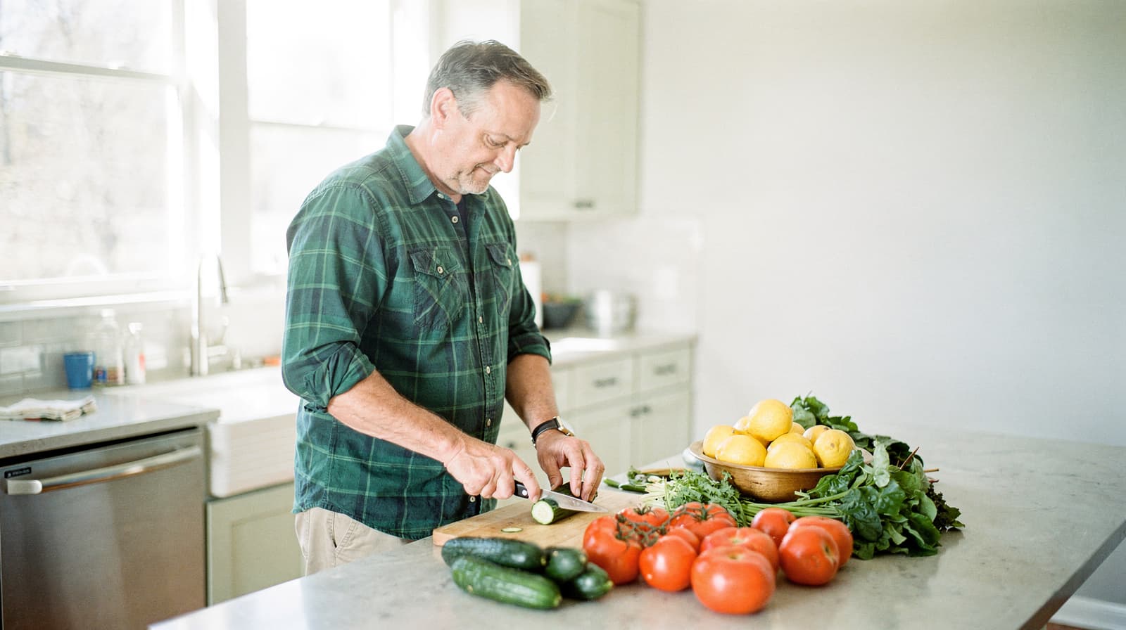 A man in a green shirt slices a cucumber on a kitchen counter with various vegetables around him.
