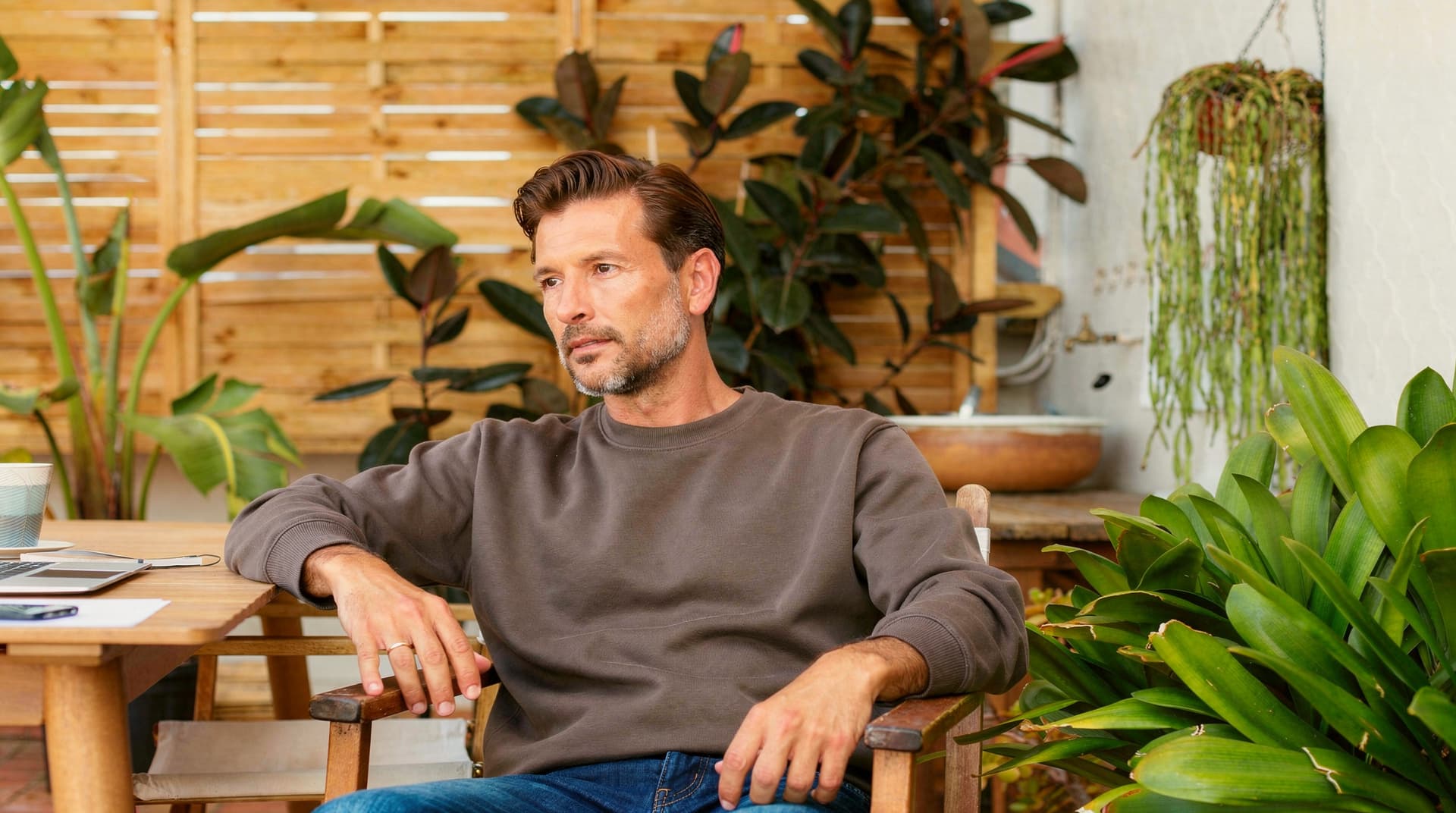 A man sits on a wooden chair in a garden patio surrounded by plants.