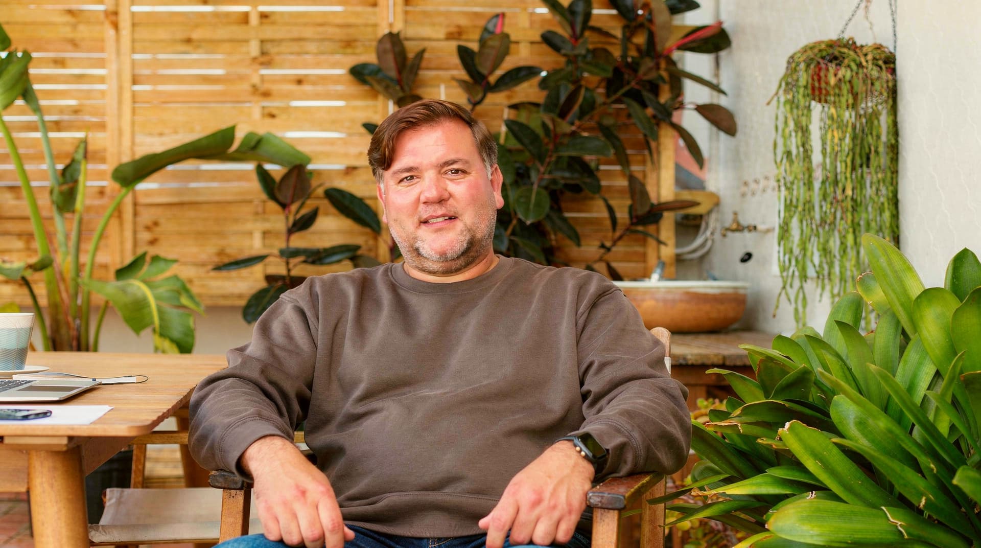 A man sits on a wooden chair in a garden patio surrounded by plants.