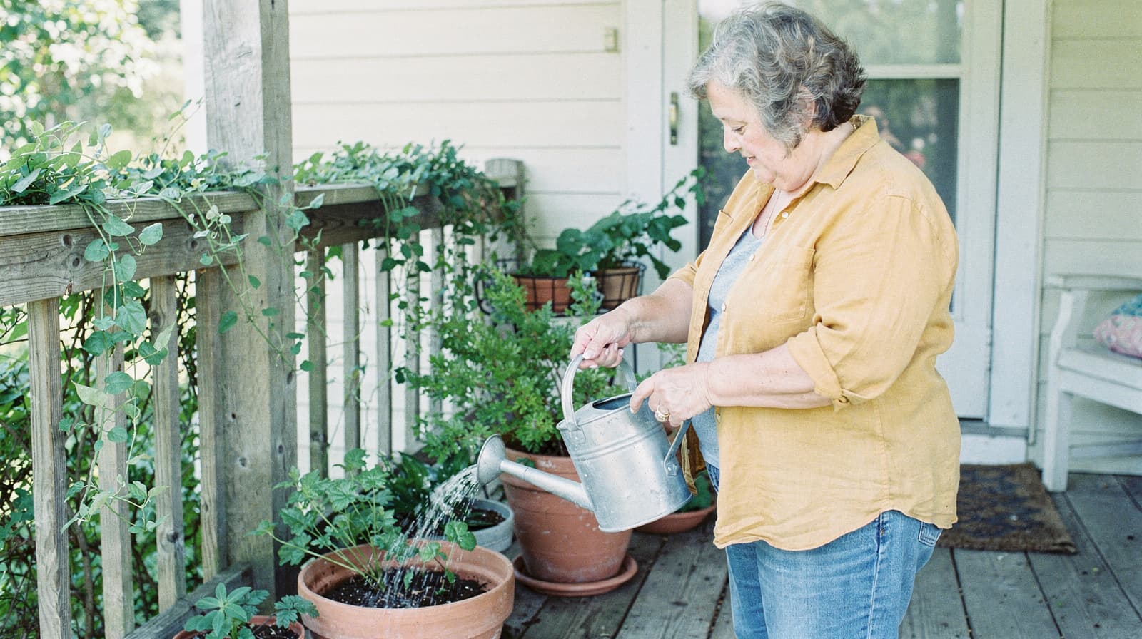A woman waters plants in pots on a wooden porch next to a railing with vines.