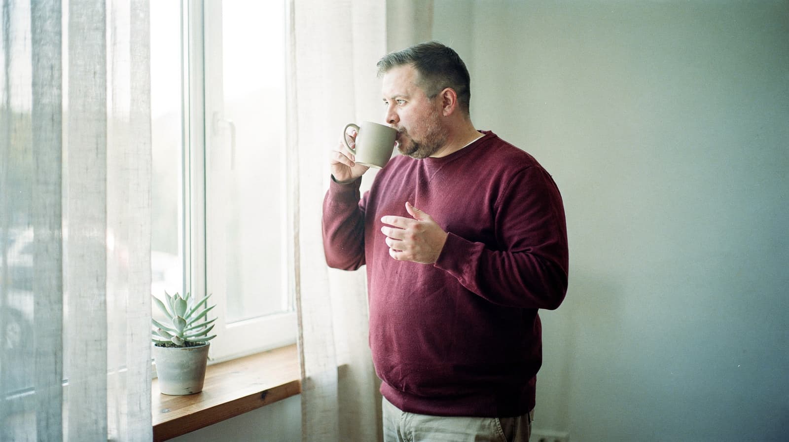 A man in a red sweater stands by a window drinking from a mug, with a small potted plant on the windowsill.
