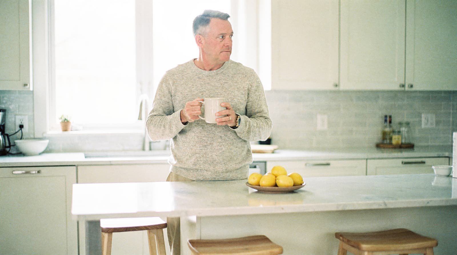 A man holding a mug stands in a bright kitchen next to a counter with a bowl of lemons.