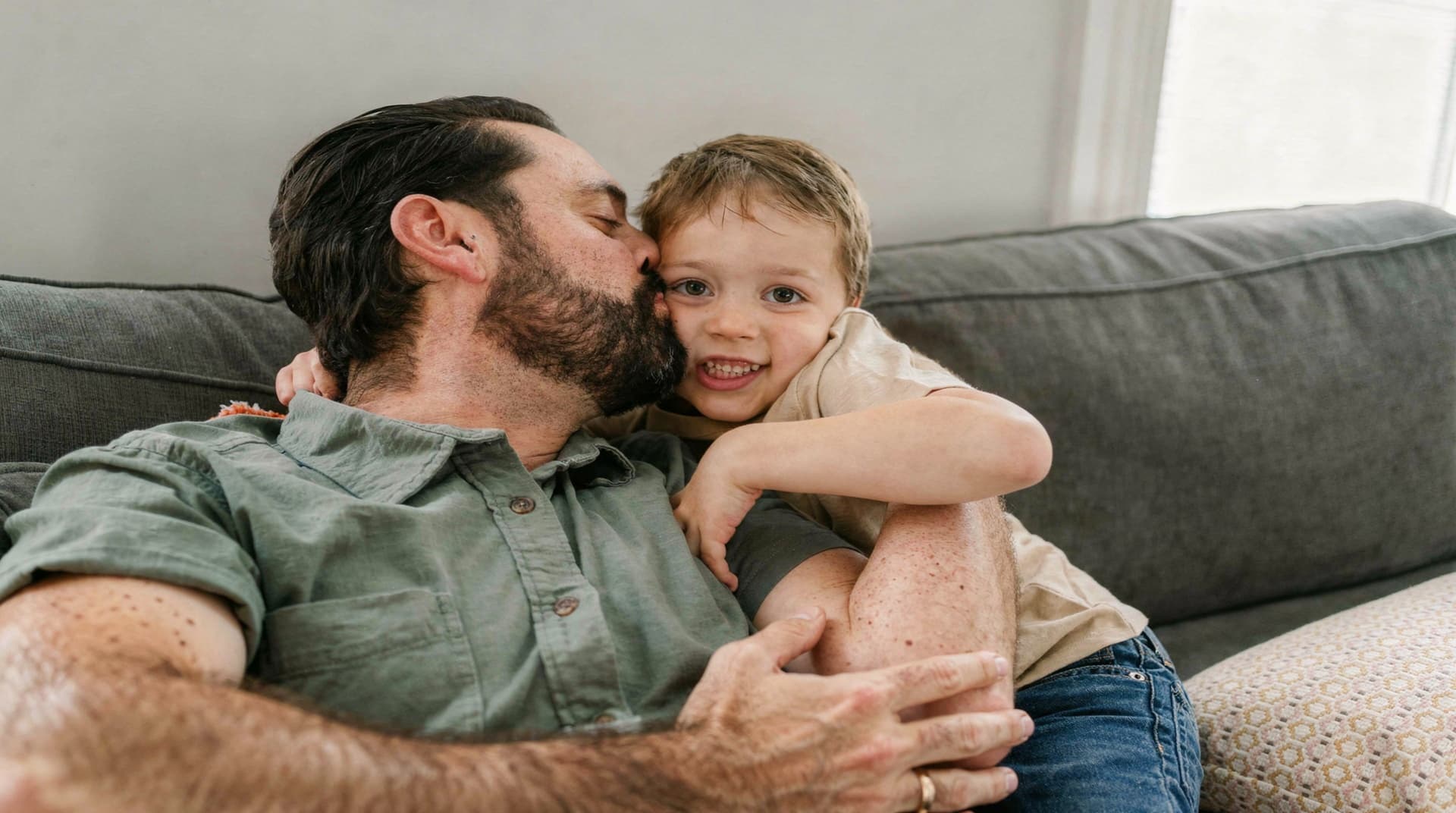 A man with a beard kisses a smiling child on the cheek while sitting on a gray couch.