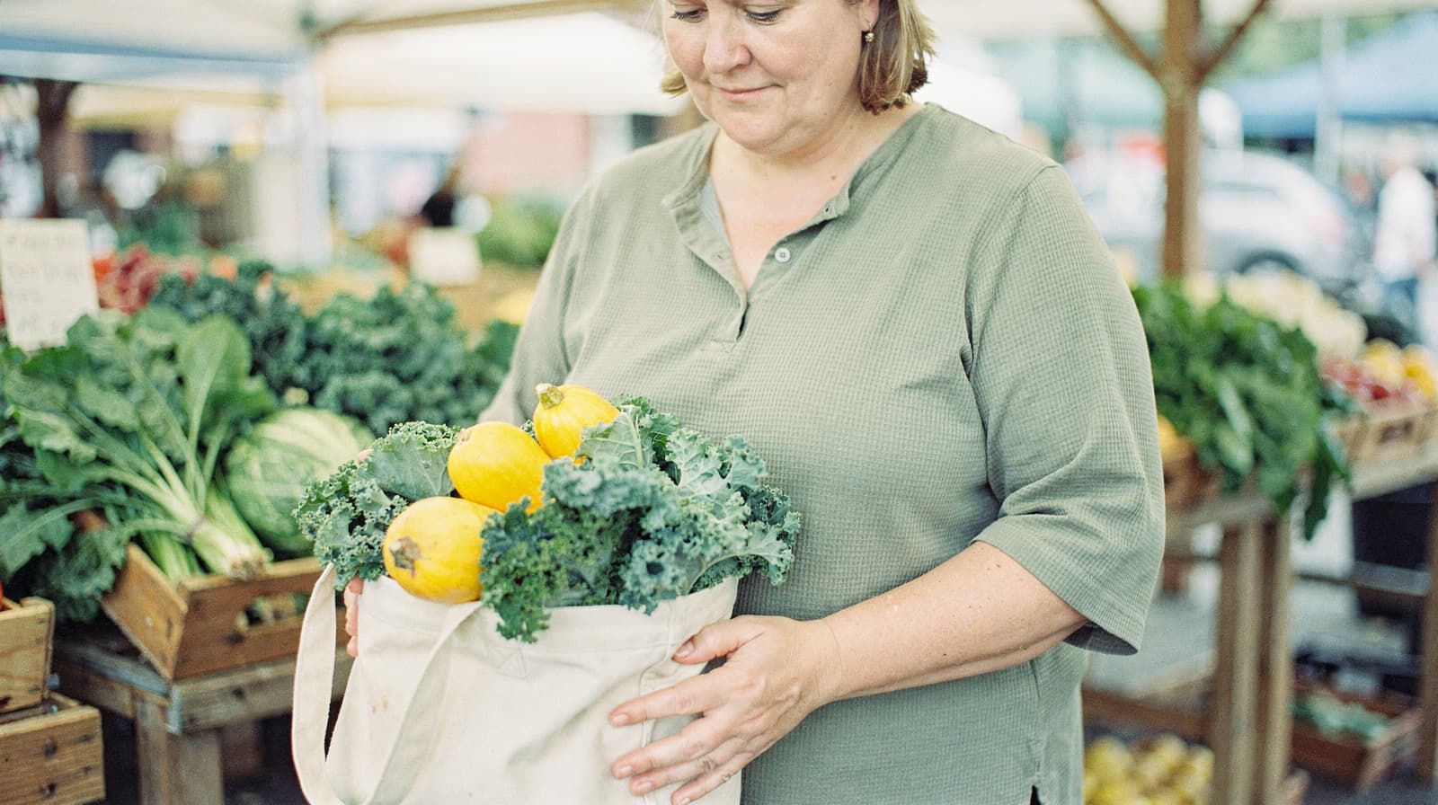 A woman holds a bag of fresh produce, including kale and squash, at an outdoor market.