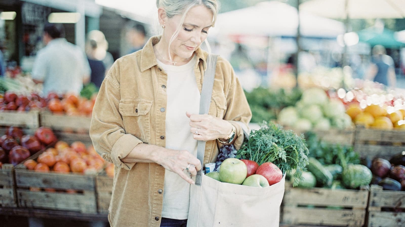 A woman stands at an outdoor market holding a bag filled with fresh produce.