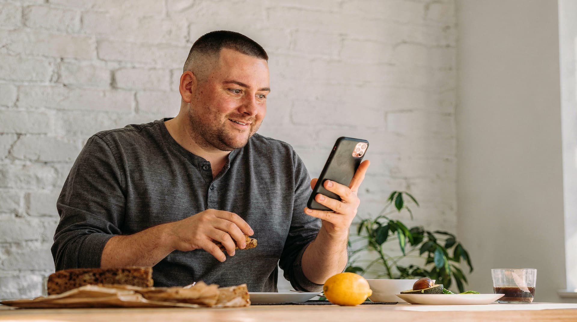 A man sits at a table with food, holding a phone and smiling, in a room with a white brick wall and a plant.