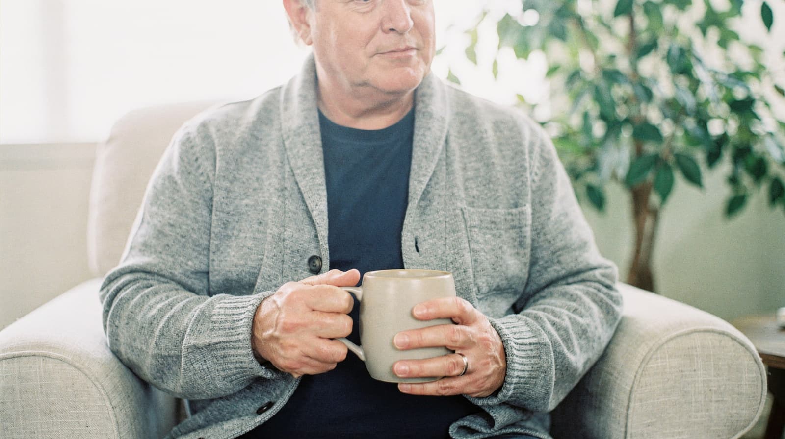 A man sits in an armchair holding a mug, with a plant in the background.