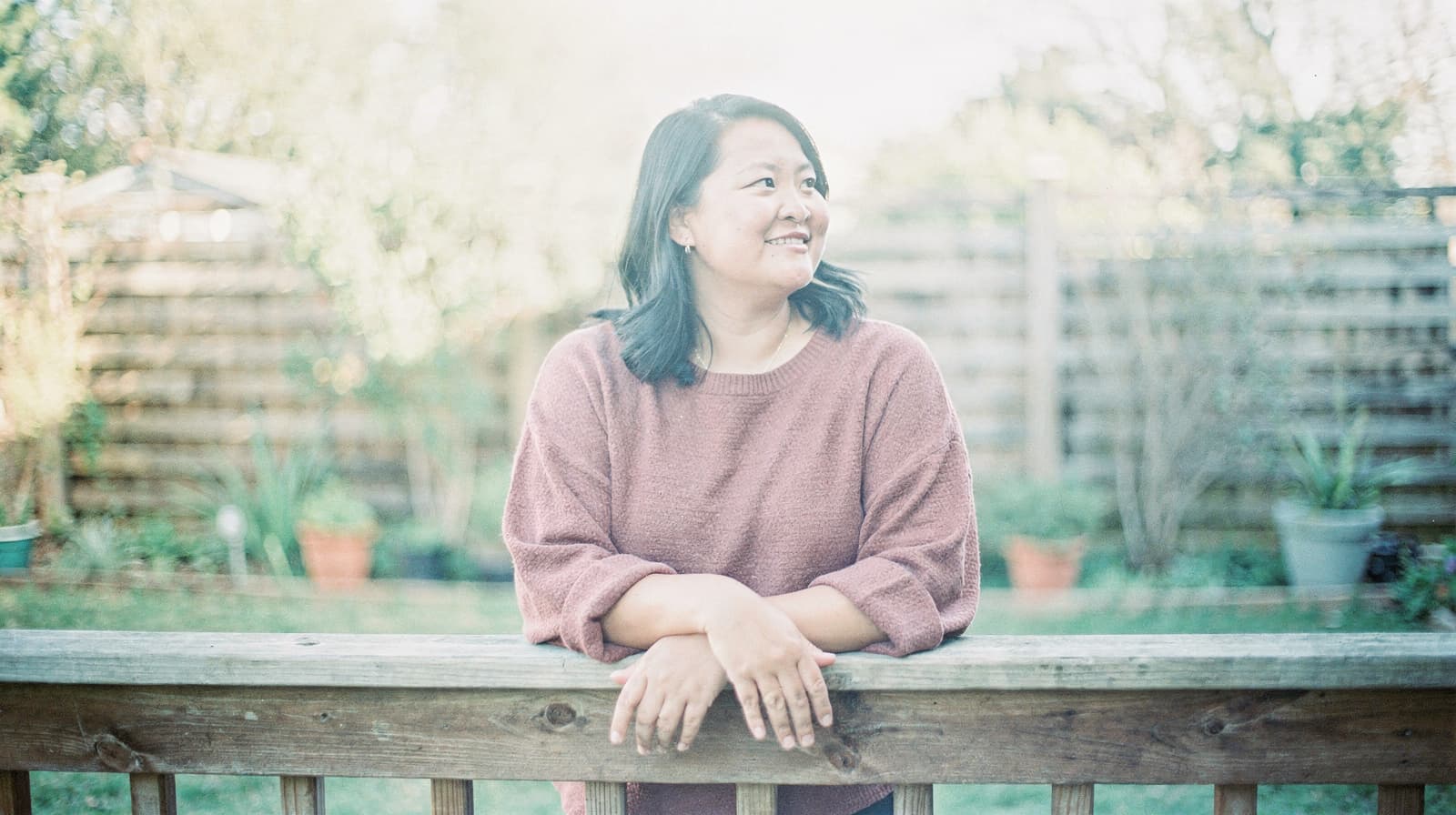 A woman in a pink sweater leans on a wooden fence in a garden, smiling and looking to the side.