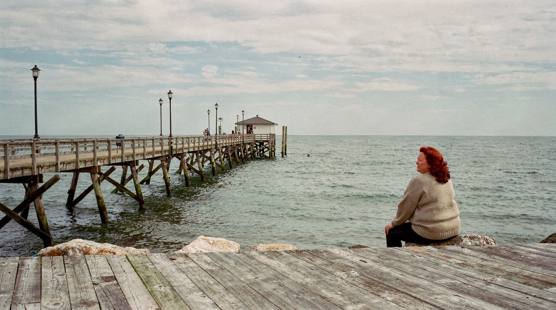 A woman sits on a rock near a wooden pier extending into the ocean under a cloudy sky.