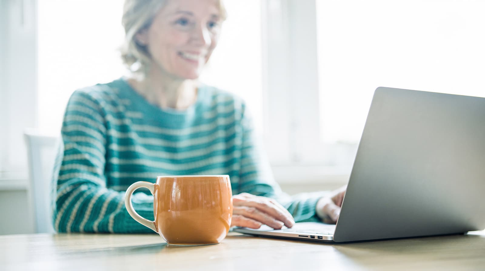A woman in a striped sweater uses a laptop at a table with an orange mug in front of her.